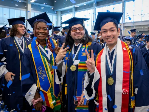 Graduates in caps and gowns pose and smile while confetti falls.