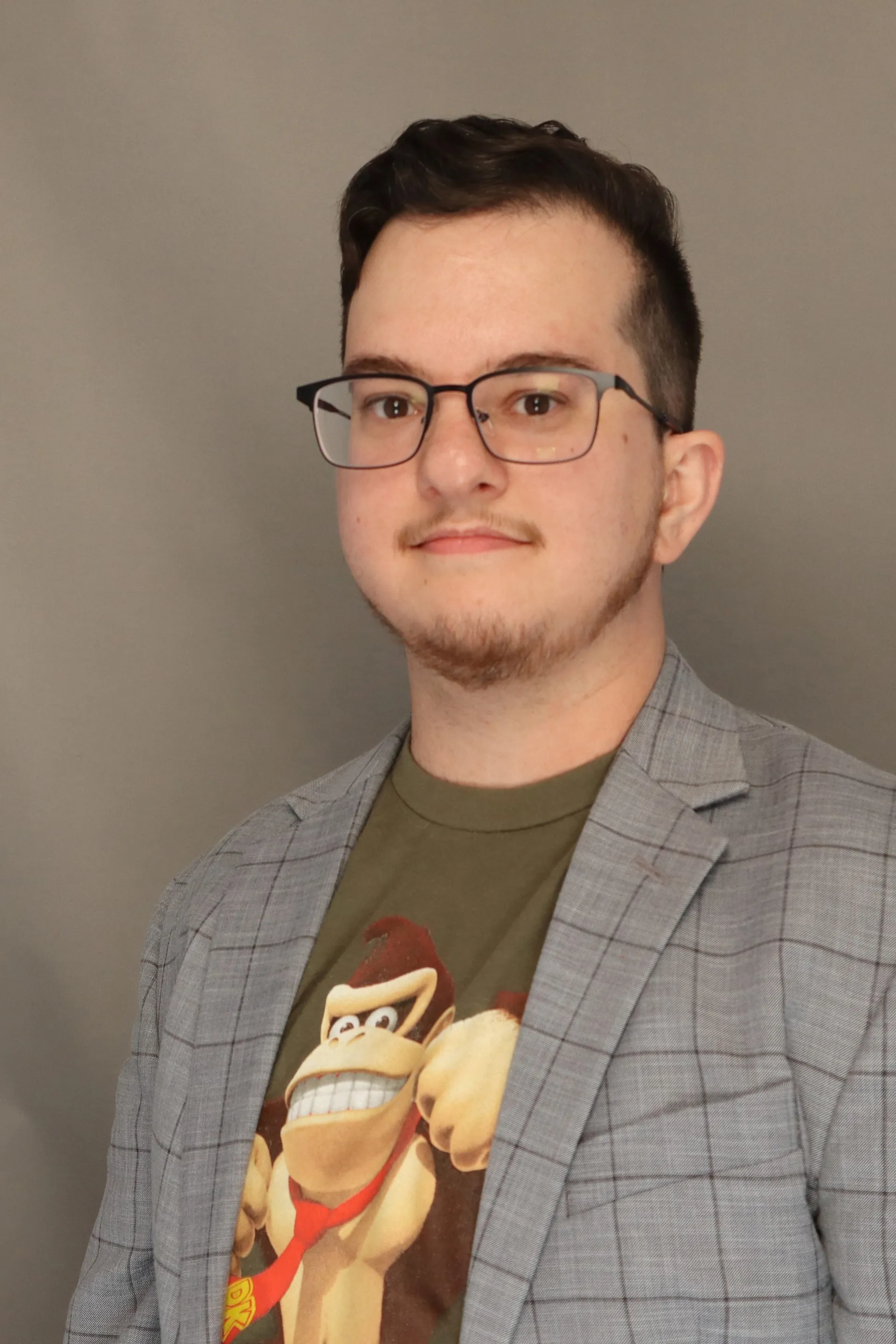 A headshot of a Caucasian man in his early 30s wearing glasses, a grey jacket, and a t-shirt with a graphic of Donkey Kong on it.