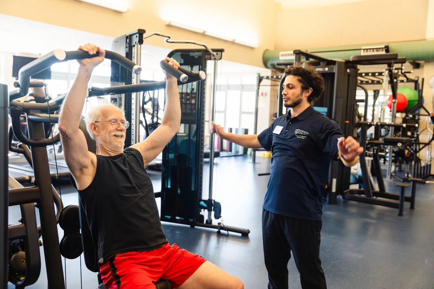 A college student assists a 65+ community member in a fitness facility.