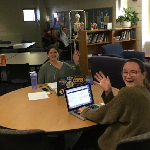 A large room with couches and worktables. Three students are seated with work at the tables and are smiling and waving at the camera